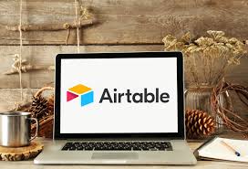**Alt Text:** A laptop on a rustic wooden desk displaying the Airtable logo, surrounded by pinecones, dried flowers, a metal mug, and a notebook.