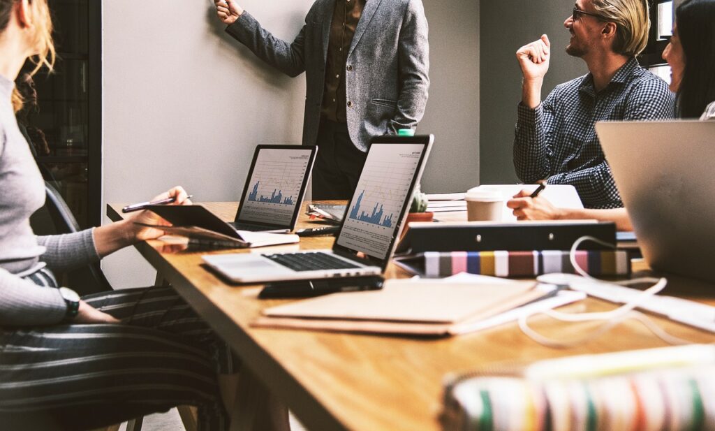 **Alt text:** A group of people in a meeting room, with laptops displaying graphs on a table, while one person points at a whiteboard.