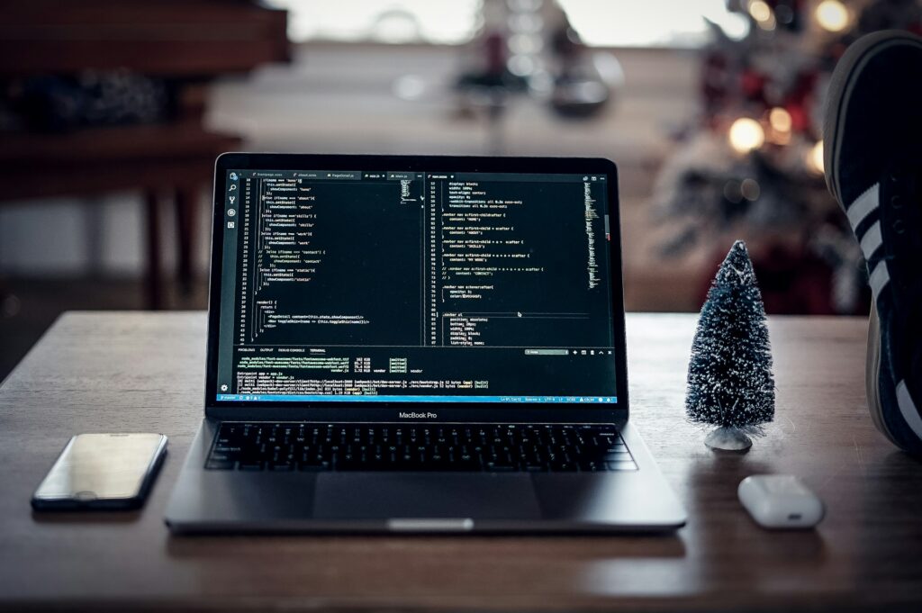 A laptop displaying code is on a table beside a small decorative tree, a smartphone, and wireless earbuds, with a blurred festive background.
