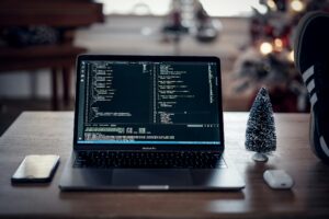 A laptop displaying code is on a table beside a small decorative tree, a smartphone, and wireless earbuds, with a blurred festive background.