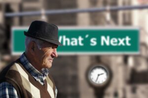 An elderly man wearing a hat and patterned sweater stands in profile with a blurred sign behind him that reads "What's Next."
