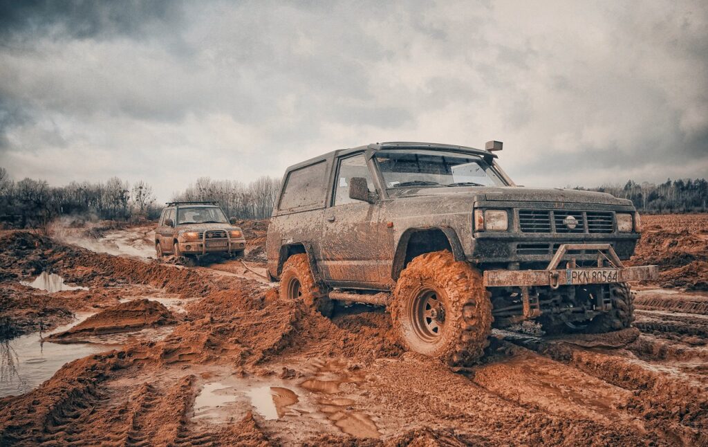 Two muddy off-road vehicles driving through a challenging, wet, and rugged terrain.