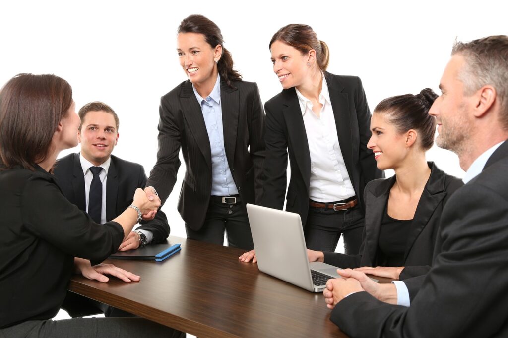 A group of business professionals in suits are sitting around a table, with two women shaking hands and smiling.