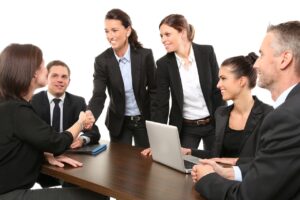 A group of business professionals in suits are sitting around a table, with two women shaking hands and smiling.