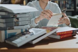 **Alt text:** A person is sitting at a table surrounded by stacks of books, reading from one in their hands.
