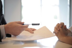 A person in a suit hands a pen and document to another person with clasped hands.