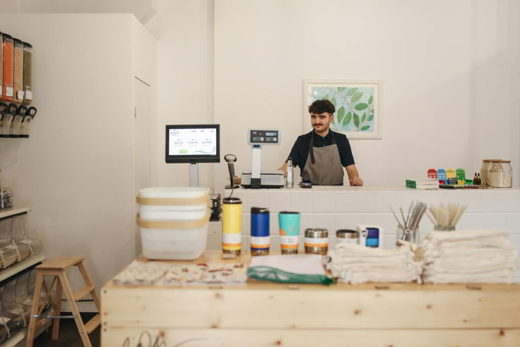 **Alt Text:** A man wearing an apron stands behind the counter of a zero-waste store, surrounded by containers and eco-friendly products.