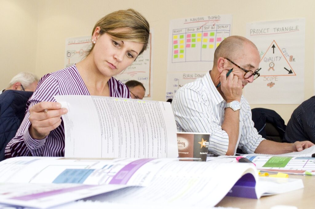 **Alt Text:** A woman and a man are focused on reading documents at a table, with charts and notes on the wall behind them.