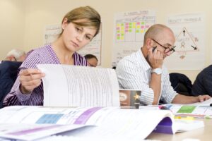 **Alt Text:** A woman and a man are focused on reading documents at a table, with charts and notes on the wall behind them.