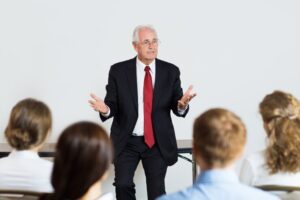 An older man in a suit and red tie speaks to a group of people seated in front of him.