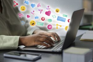 A person typing on a laptop with various colorful social media icons floating around the screen.