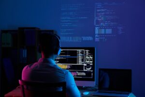 A person wearing headphones sits in front of a computer monitor displaying code, with additional code projected on a wall in a dimly lit room.