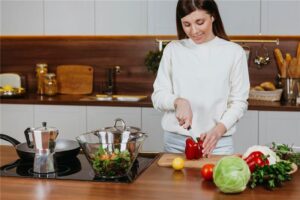 A woman in a white sweater is cutting a red bell pepper on a wooden cutting board in a modern kitchen surrounded by various vegetables.