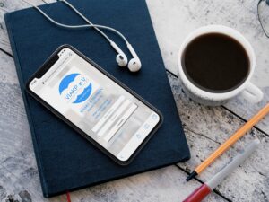 **Alt Text:** A smartphone displaying a login screen sits on a closed notebook beside a cup of coffee, earphones, a pen, and a pencil on a wooden surface.