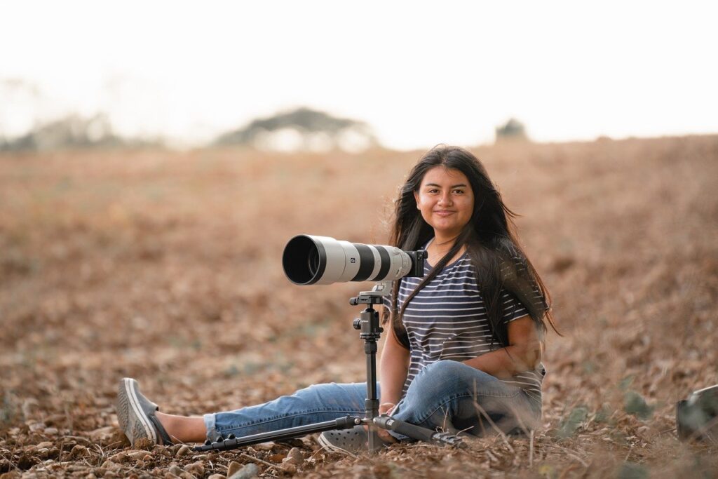 A woman sitting on the ground in a field, smiling, with a camera on a tripod beside her.