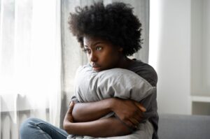 A person with curly hair sits on a couch holding a pillow, looking pensively out of a window.