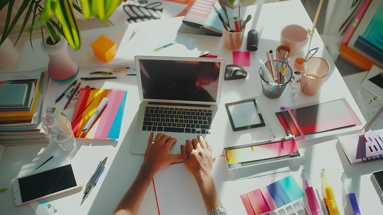 A person working at a cluttered desk filled with colorful stationery, a laptop, a smartphone, and various office supplies.