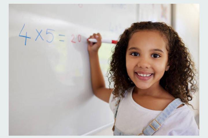 A young girl smiles while holding a marker in front of a whiteboard displaying the equation "4 x 5 = 20."