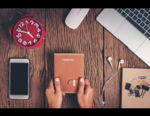A person holds a brown passport cover on a wooden desk surrounded by a red alarm clock, smartphone, mouse, laptop, earphones, and a notebook with paper clips.