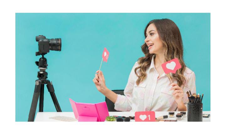 A woman sitting at a table with makeup products, holding heart-shaped social media icons and smiling at a camera on a tripod.