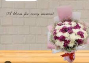 A bouquet of pink and white flowers is placed on a wooden surface with a brick wall backdrop and the words "A bloom for every moment" written above it.