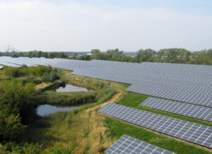 **Alt Text:** A large solar farm with rows of solar panels surrounded by greenery and a small pond.
