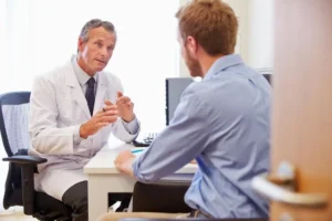 A doctor in a white coat is sitting at a desk, attentively listening to a patient seated across from him.