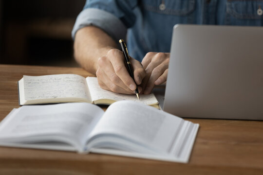 **Alt text:** A person is writing in a notebook while working on a laptop, with an open book in the foreground on a wooden desk.