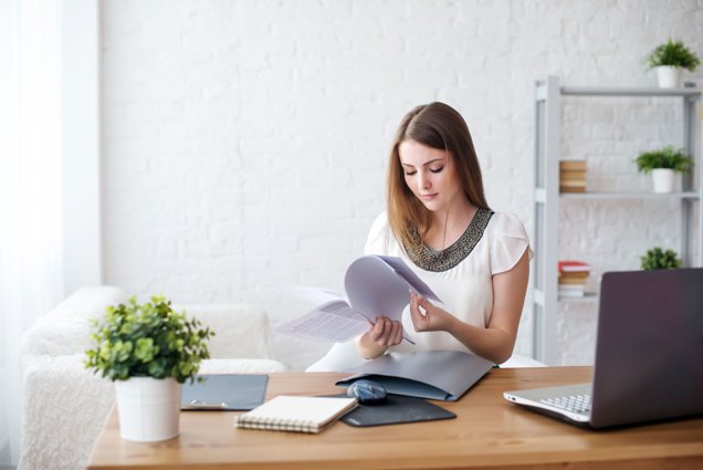 **Alt Text:** A woman sits at a desk reviewing documents with a laptop, plant, and notebooks nearby in a bright room.