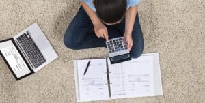 **Alt text:** A person sits cross-legged on a carpet, using a calculator while referencing documents in a binder and a laptop placed nearby.