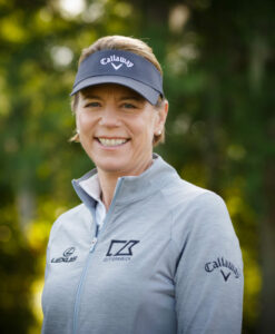 **Alt text:** A person wearing a grey sports jacket with sponsor logos and a dark visor smiles outdoors with a blurred green background.