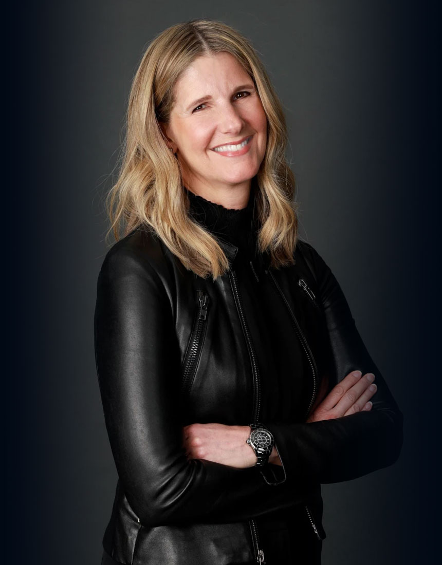 Alt text: A woman with shoulder-length blonde hair smiles, wearing a black leather jacket and a watch, with arms crossed.