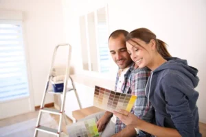 Alt text: A couple is sitting together, smiling while looking at a paint color swatch, with a ladder and painting supplies in the background.