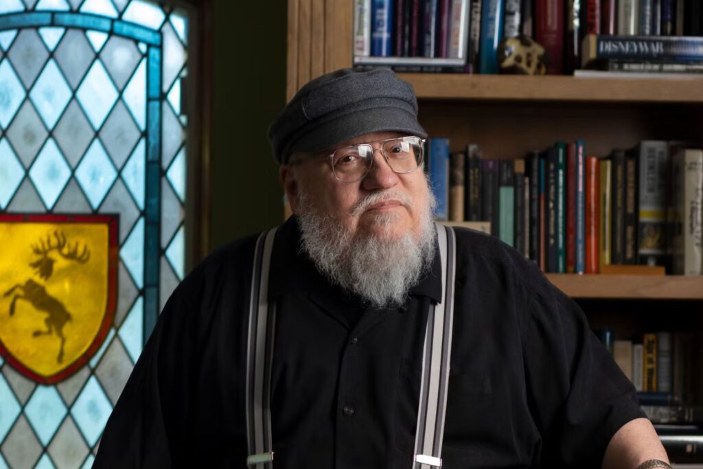 Alt text: A person with a white beard and wearing a cap and suspenders stands in front of a bookshelf, with a stained glass window featuring a stag in the background.