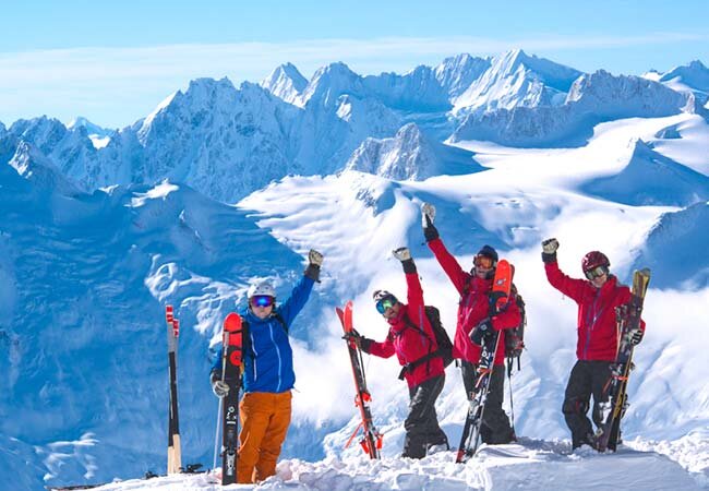 **Alt Text:** Four skiers in colorful outfits stand triumphantly on a snowy mountain peak with skis and poles, set against a backdrop of snow-covered mountains.