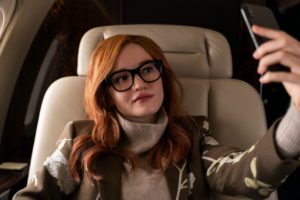 Alt text: A woman with red hair and glasses takes a selfie while sitting in an airplane seat.