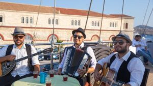 **Alt Text:** Three musicians wearing hats and sunglasses play instruments on a boat deck in front of a historic building, with cups of tea on the table.