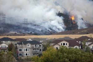 **Alt text:** A wildfire with billowing smoke and visible flames burns on a hill near a residential area with houses in the foreground.