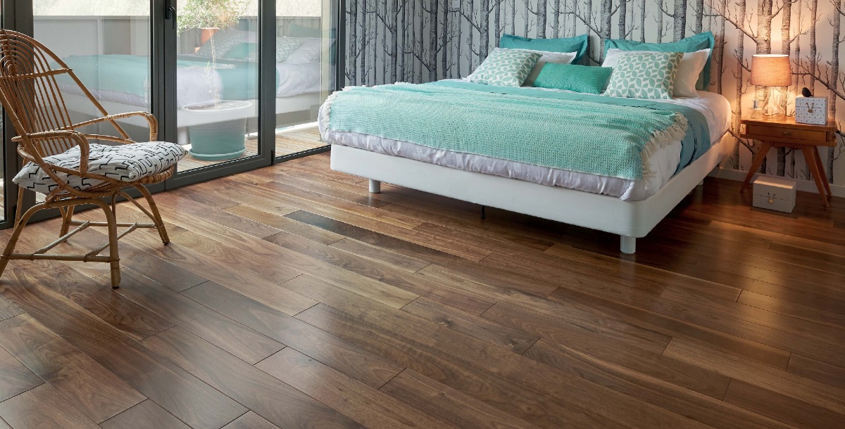 **Alt text:** A modern bedroom with dark wooden floors, a bed with teal and white bedding, a patterned wallpaper, a wooden chair, and a bedside lamp on a small table.