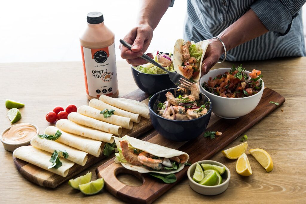 A person is preparing tacos on a wooden board with ingredients including tortillas, chipotle mayo, vegetables, and lime wedges.