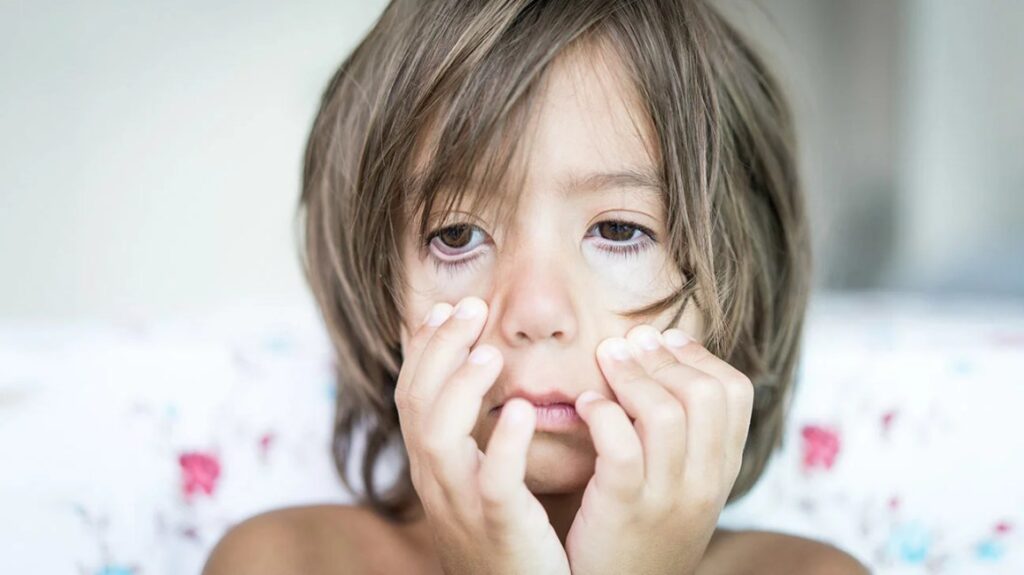 A child with long hair holds their face with both hands, looking tired or pensive.