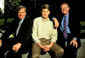 Alt text: Three men in formal and casual attire sit together outside on a bench.