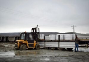 A forklift loads large concrete blocks onto a flatbed truck while a person adjusts cargo straps under a cloudy sky.