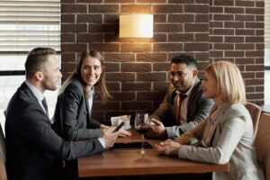 <!-- Four people in business attire are sitting around a wooden table in a brick-walled restaurant, engaged in conversation with a glass of wine visible on the table. -->