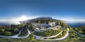 **Alt text:** A panoramic aerial view of a large, historic estate surrounded by lush gardens and coastline, with mountains in the background under a clear blue sky.