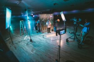 A brightly lit film set featuring a person standing in front of a glittery backdrop, surrounded by lighting equipment and a camera crew in a large indoor space.