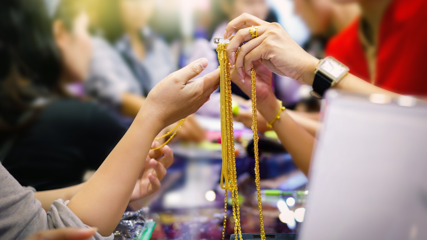 **Alt Text:** A person is holding up several gold necklaces while another person examines them in a jewelry store.