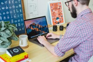 **Alt Text:** A bearded man wearing a plaid shirt is working on a laptop at a desk, surrounded by books, a plant, and camera lenses, with a motivational poster on the wall.
