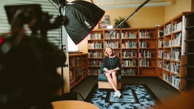 **Alt text:** A woman is sitting on a chair in a library, surrounded by shelves of books, with professional lighting and camera equipment set up for an interview.
