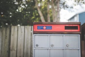 **Alt Text:** A Canada Post community mailbox stands against a wooden fence with trees and a house in the background.
